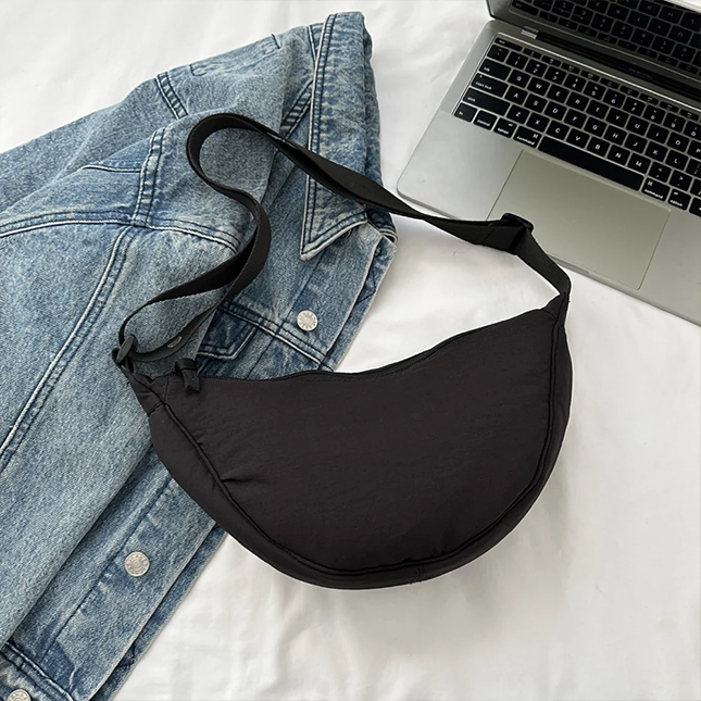 Half moon crossbody sling bag in black laid flat beside a denim jacket and laptop