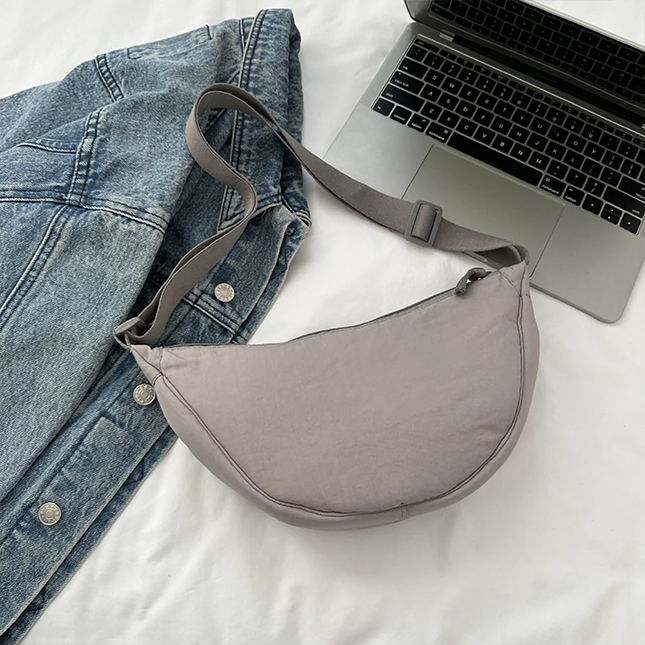 Half moon crossbody sling bag in grey laid flat beside a denim jacket and laptop